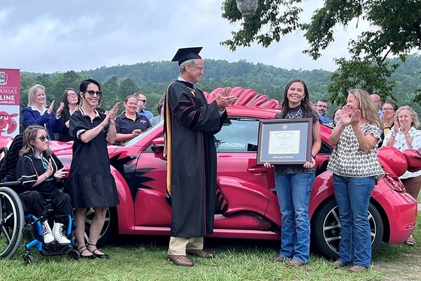 student stand with degree in front of Razorbug car and people clapping