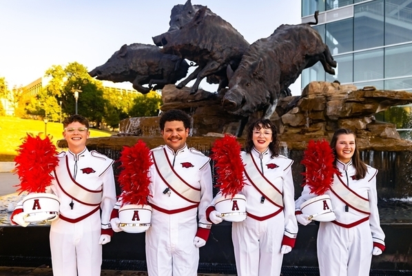 Drum majors are lined up holding their hats in front of a Razorback statue