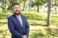 Seamus O'Brien wearing a blue sport coat and a pink shirt stands outdoors on SUNY Canton's campus. 