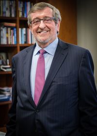 Man smiling, wearing a business suit and glasses in front of a bookshelf background. 