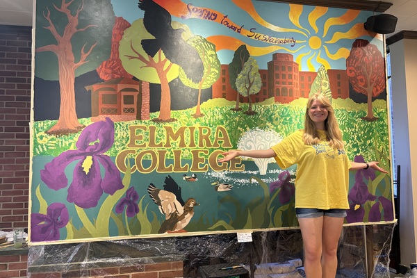 A woman with blond hair in a yellow T-shirt and jean shorts stands in front of a colorful mural depicting some of the campus grounds with wildlife. 