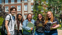 A group of five students smiling in front of a university building holding laptops and books