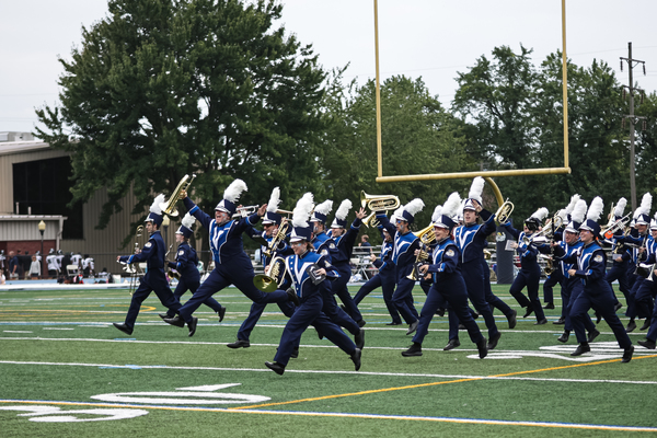 Marching band members in uniform run onto the football field with their instruments.