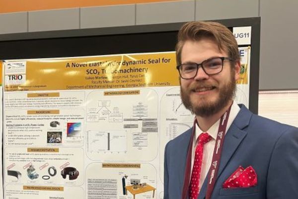 Man with glasses and a beard in a blue suit and red tie stands beside an academic poster at a conference. 
