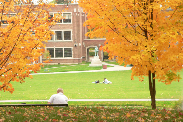 Several students are seated. In the foreground, two trees have bright yellow-orange leaves. In the background are academic buildings.