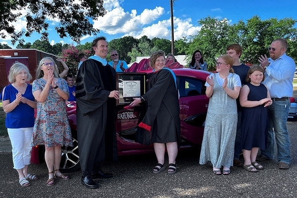 graduate in cap and gown accepts diploma with family standing around clapping. 