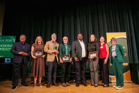Seven SUNY Delhi alumni award winners pose on stage with their trophies and plaques next to President Mary Bonderoff.