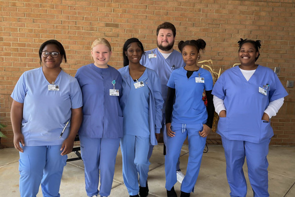Zoey Jones, Josey Stevens, Jamiyah Levens, Destry Hunter, Denetrius Wade, and Shuntyia Brooks stand outside a brick building in blue scrubs