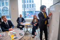 3 individuals seated wearing business attire, watching another individual doing a presentation on a whiteboard in a modern space.