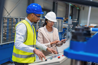 Female manager guides male worker on production process in busy factory. Focus on collaboration and workplace communication.
