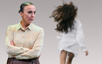 A woman standing with arms crossed in foreground with a woman running away in the background.