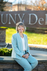 Dr. Mary Bonderoff sitting and smiling in front of the SUNY Delhi campus sign.