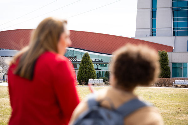 Two blurred visitors, one in a red coat and one with a backpack, face Goodwin University's main building on a bright day.