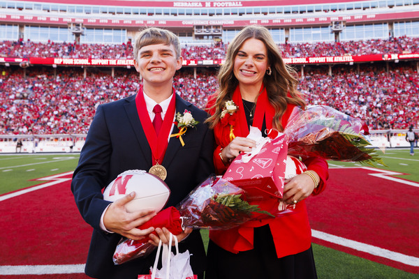 A young man and young woman in black-and-red formal attire pose with flowers and gifts at midfield in Memorial Stadium.