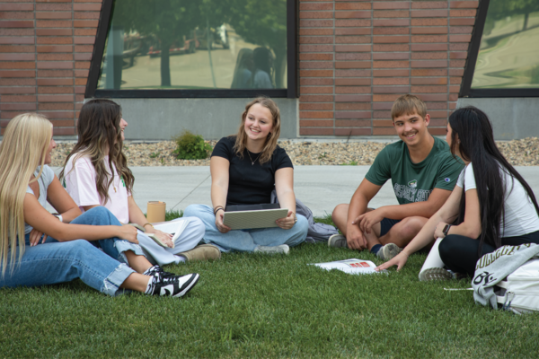 Five BSC students sit in a circle on the campus lawn, talking and studying together with books, a tablet and backpacks nearby.