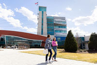 Two university students walk across a sunny campus plaza beside a modern glass-and-steel academic building, creating a welcoming mood.