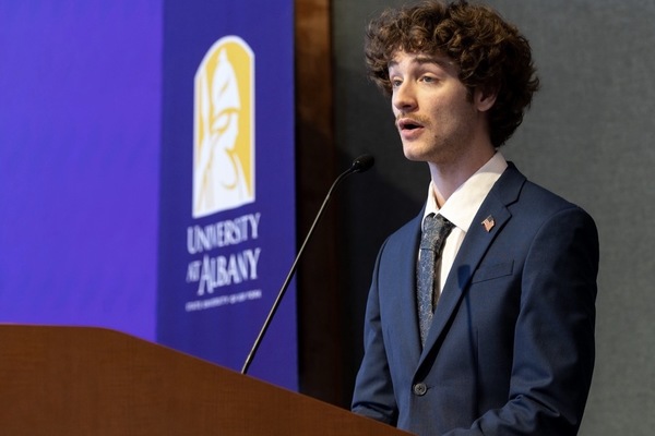 Troy Serao speaking at a podium with the University at Albany banner in the background.