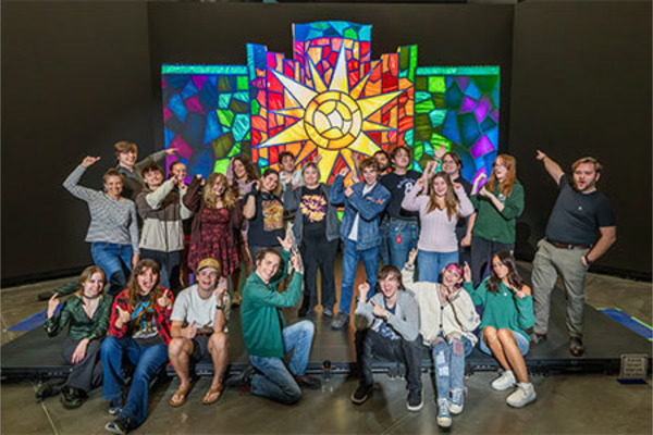 a group of students standing in front of their light display, which looks similar to a stained glass window with a sun on the center panel