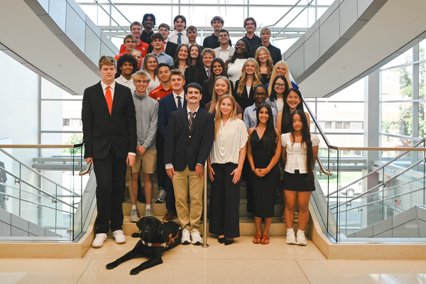 Thirty-four University of Nebraska–Lincoln students in professional attire and a black dog pose for a group photo on a staircase in Hawks Hall.