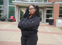 A young woman standing with her arms crossed in front of an academic building. 