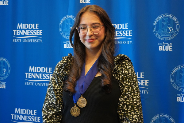 Smiling MTSU student just successfully defended Honors thesis, wearing medals in front of backdrop