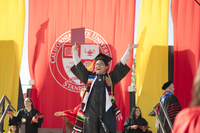 Stanislaus State graduate wearing a black cap and gown, colorful stole and honor cords raises both arms in celebration while holding a diploma cover.
