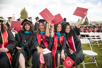 Four smiling Stanislaus State graduates in black caps and gowns sit together at commencement, with several raising red diploma covers.