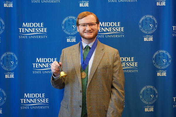 Smiling MTSU student just successfully defended Honors thesis, wearing medal and holding bell in front of backdrop