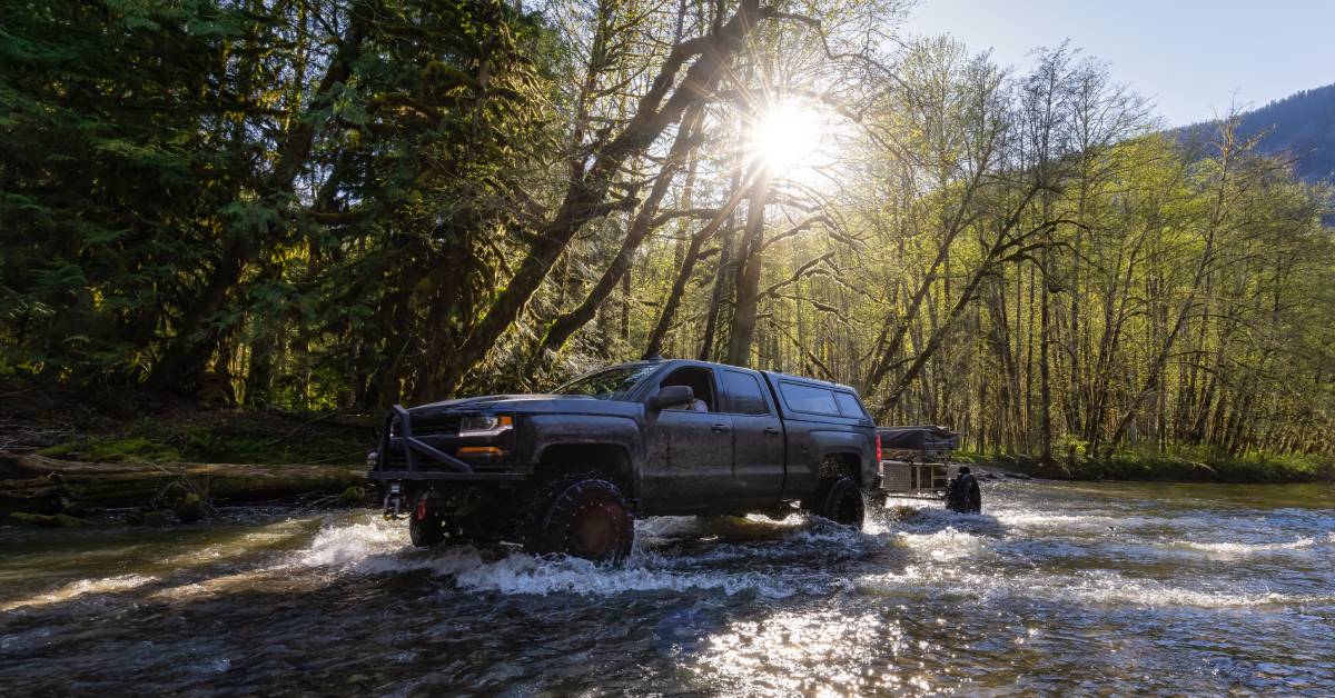 A custom pickup truck with a lifted suspension towing a trailer through a shallow stream in a backwoods forest.