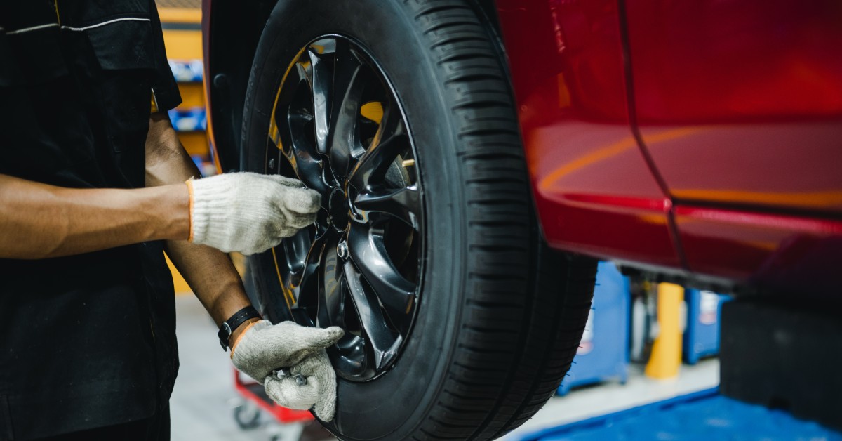 An auto mechanic wearing white gloves inspects the rim of a wheel on a lifted red car in a mechanic's garage.
