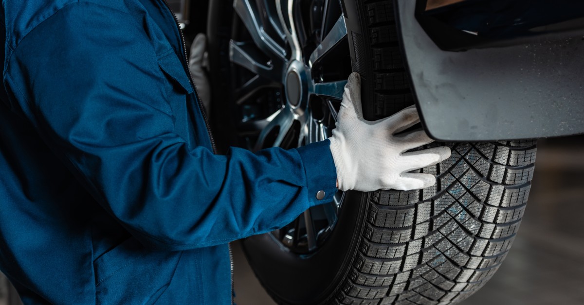 An auto mechanic in a blue jacket, pants, and white gloves holds up a new wheel and tire to a lifted car for installation.