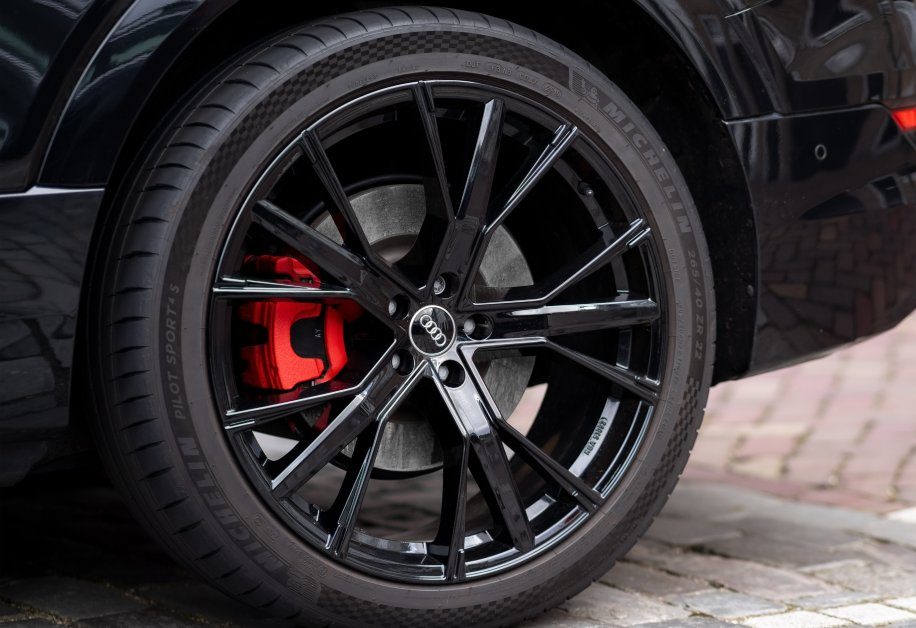 A close-up of a black Audi alloy wheel with red brake calipers behind it on a black Audi car parked on a brick street.