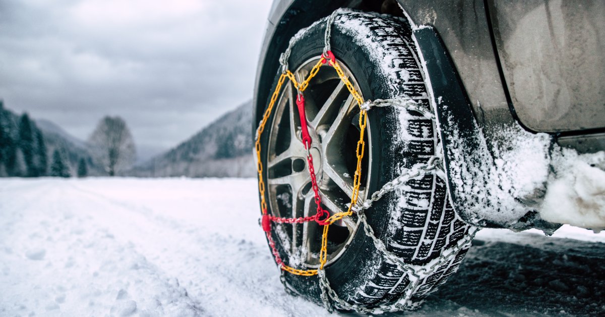 The front tire of an SUV on a snow-covered road with tire chains. Blurred in the background are mountains and forests.