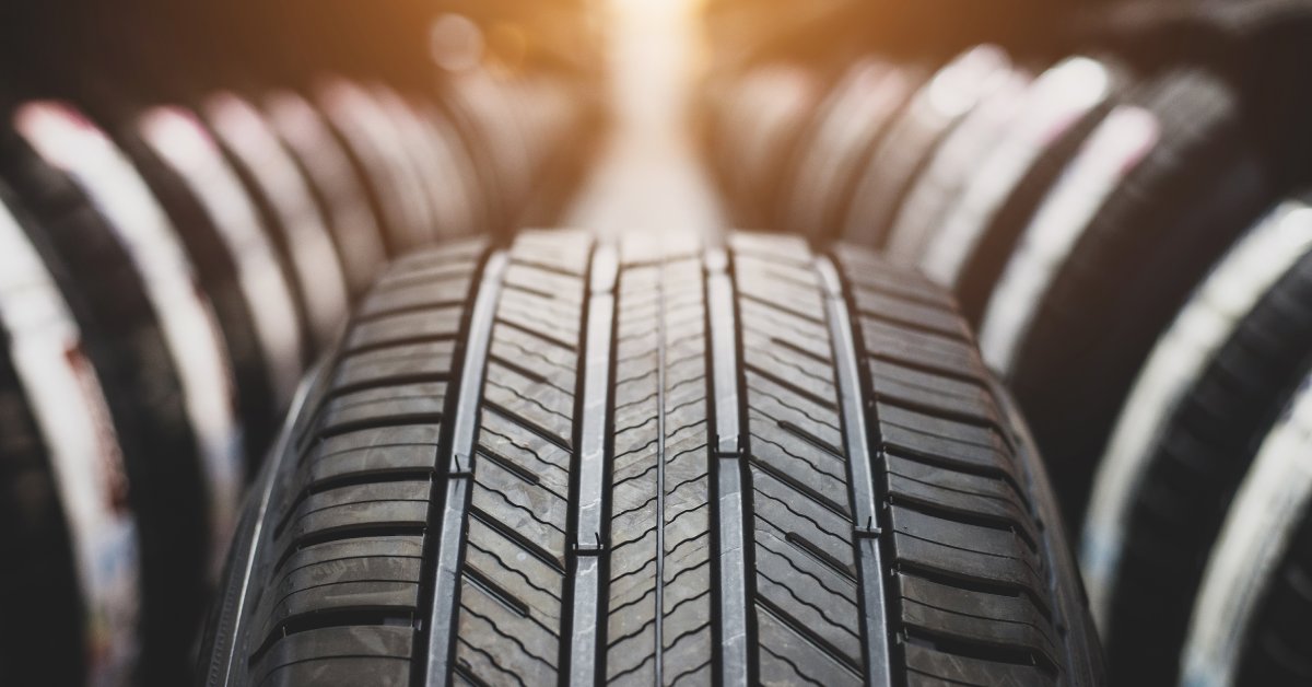 A vertical automotive tire with an asymmetrical tread pattern in focus with rows of other tires blurred in the background.