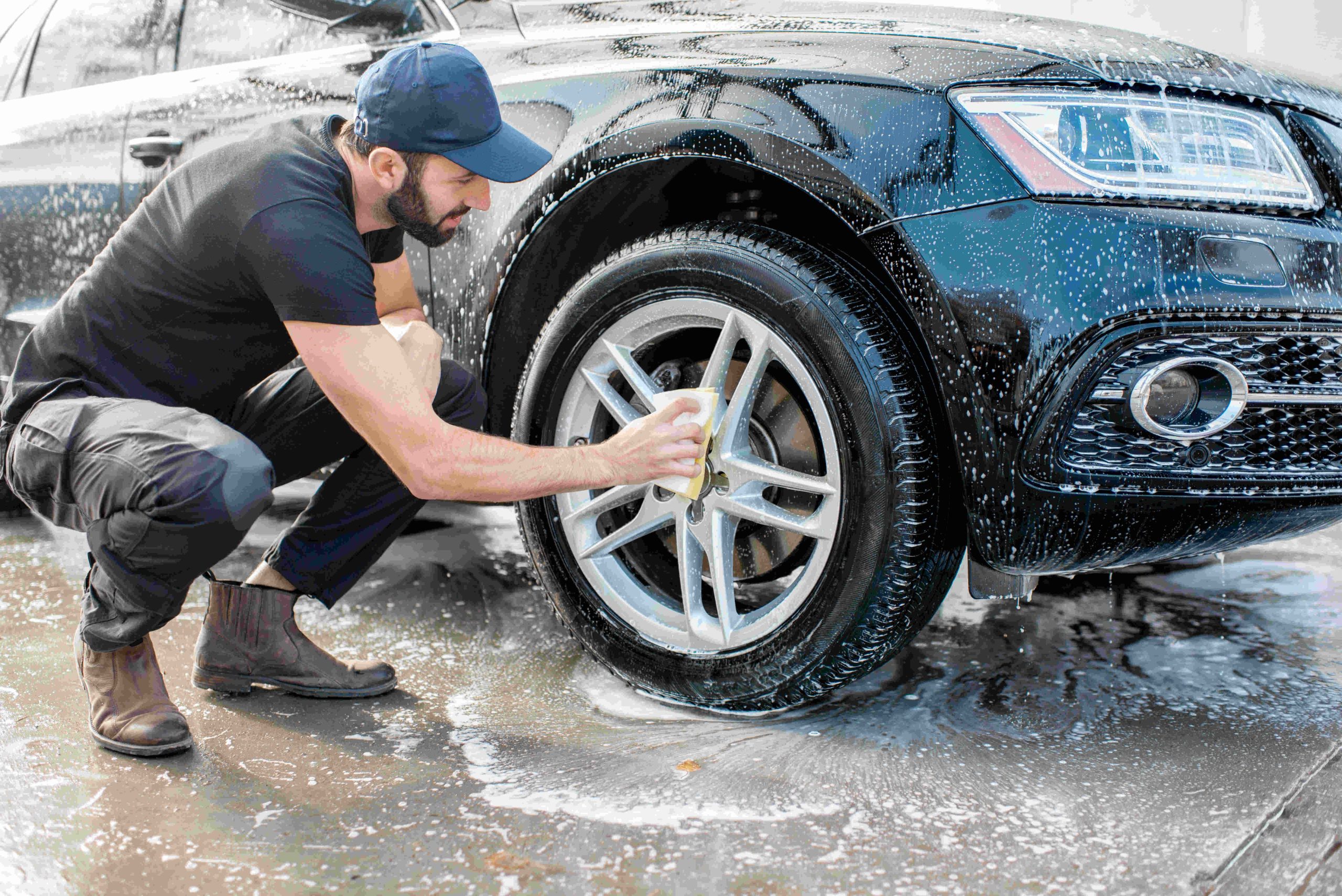 A man crouches down to scrub a wet and soapy car tire with a yellow sponge while wearing a baseball hat and boots.