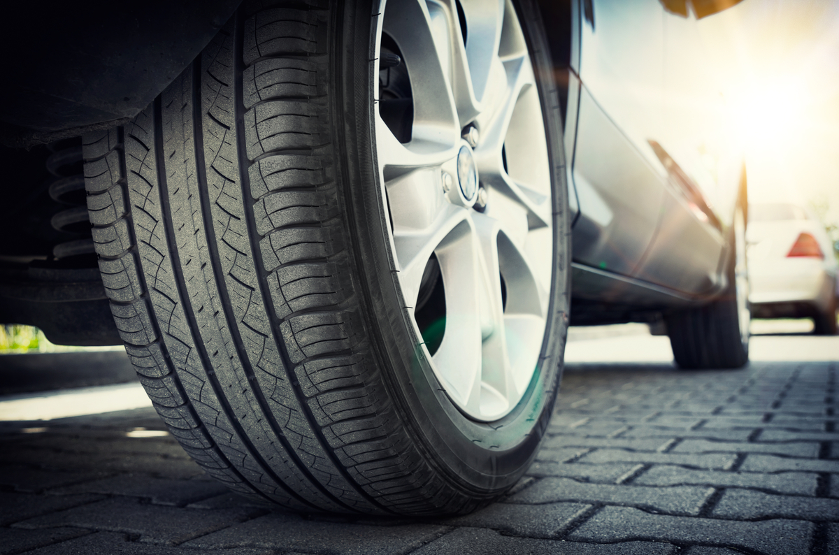 A close-up of a silver car's rear tire and wheel on a brick road with the sun glaring behind the vehicle.