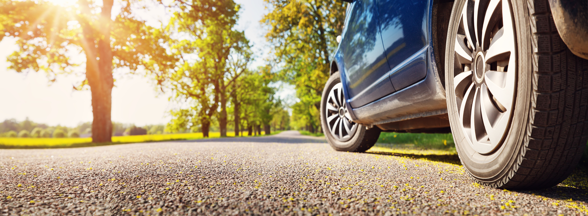 A ground-level view of a blue car and its tires on a paved road in the countryside surrounded by trees.