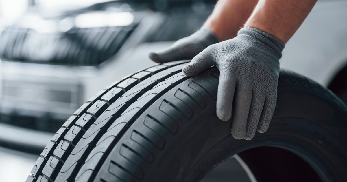 A close-up of a mechanic's hands with gray gloves pushing a new automotive tire on the ground in a garage.