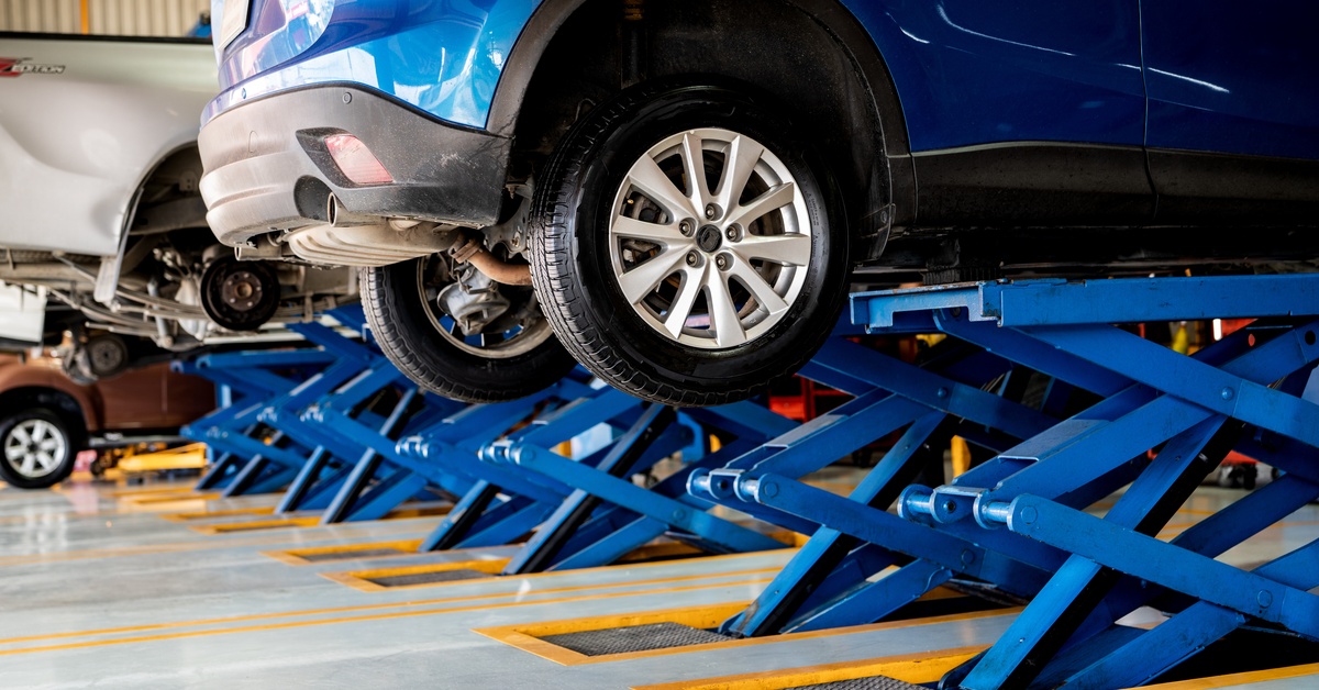 Two cars are raised in the air by blue car lifts in an auto repair shop. One vehicle is parked in the background.