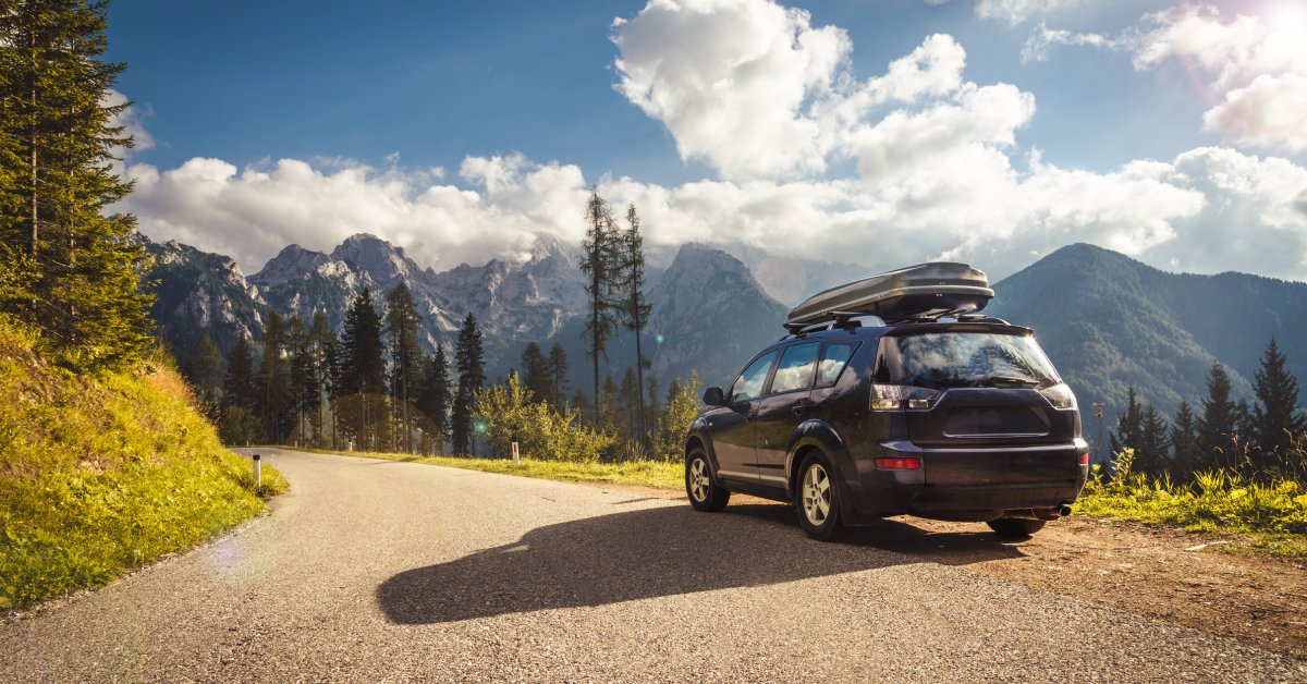 A family van with storage on the hood is traveling on a small road on a mountain, heading toward a larger mountain.