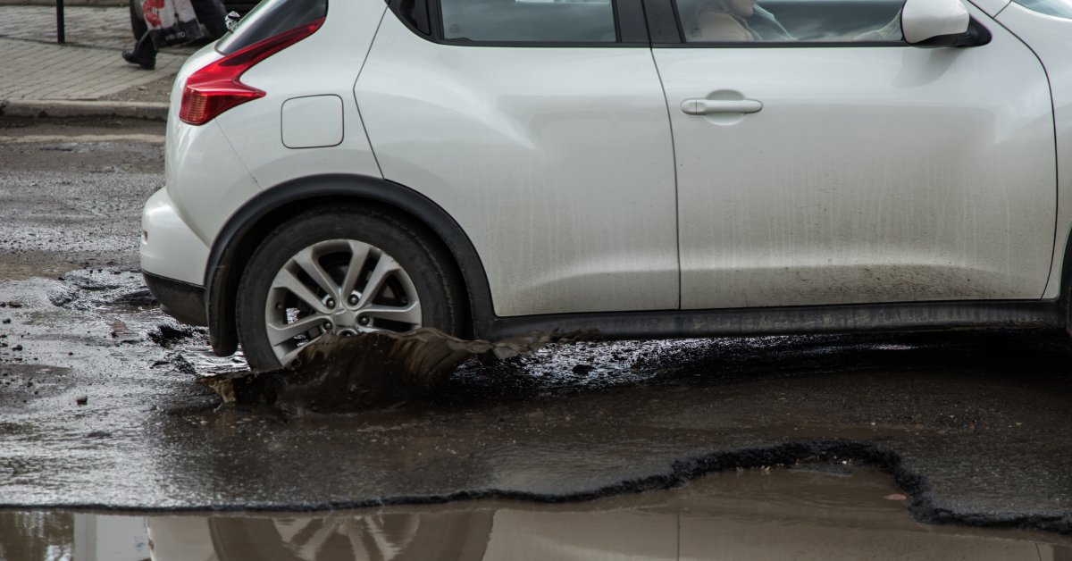 A close-up of a silver car running over a pothole. There's a massive pothole next to the pothole the car is riding over.