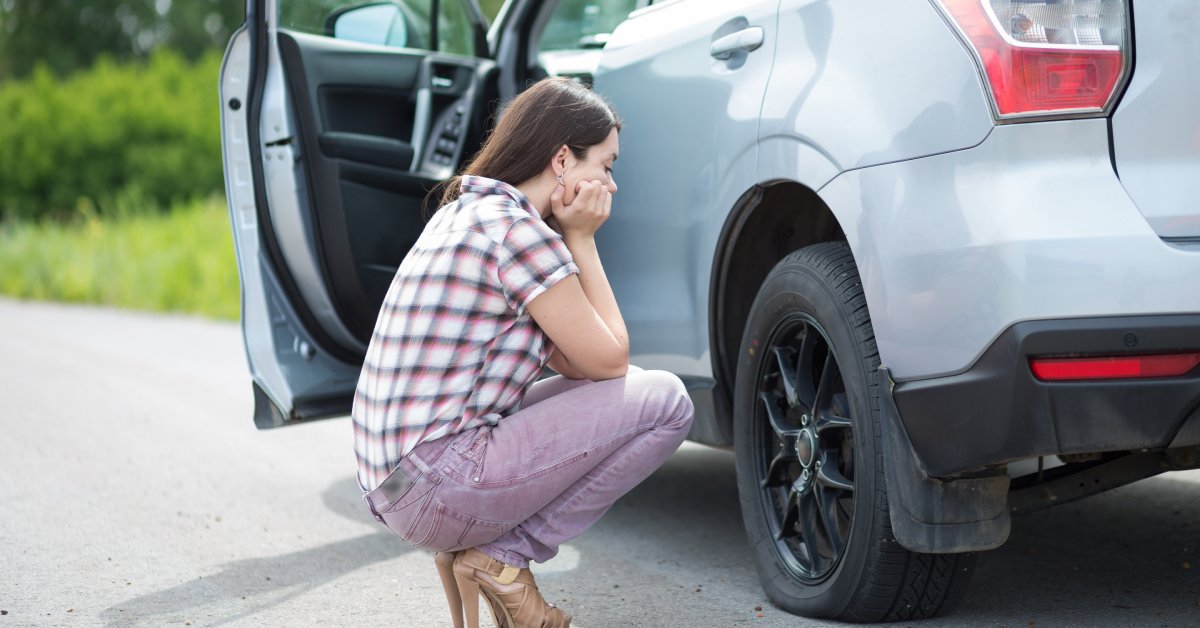 A woman wearing a plaid shirt, lilac pants, and high heels squats next to a silver SUV to inspect a flat tire.