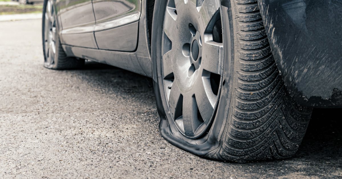 A gray car sits on the pavement with both the front and rear tires completely flat due to a puncture.
