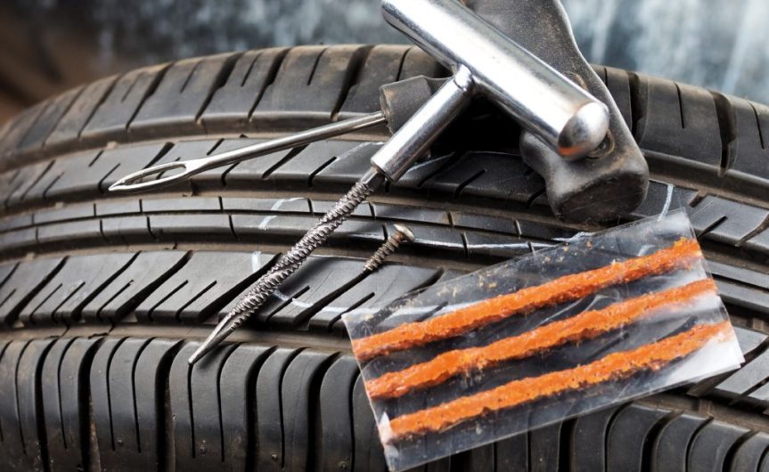 A tire with a flat tire repair kit lying on top of it. Some of the tools are a plugger, a pressure gauge, and a plug.