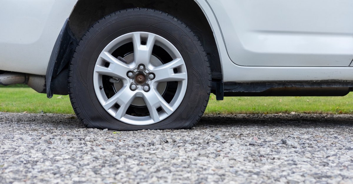 The lower half of a white car with a flat tire on the side of a gravel road. There's grass on the other side of the gravel road.