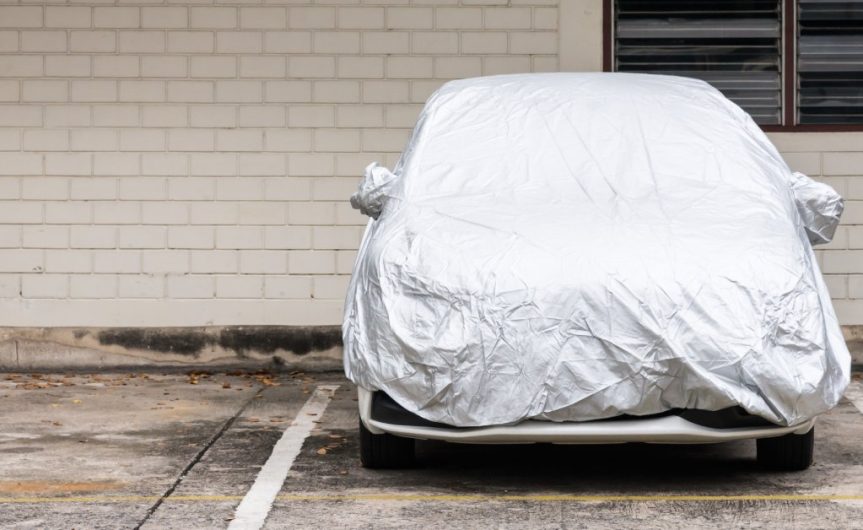 Outside of a building with wooden bricks, a car is in a parking spot. A silver cover covers the vehicle.