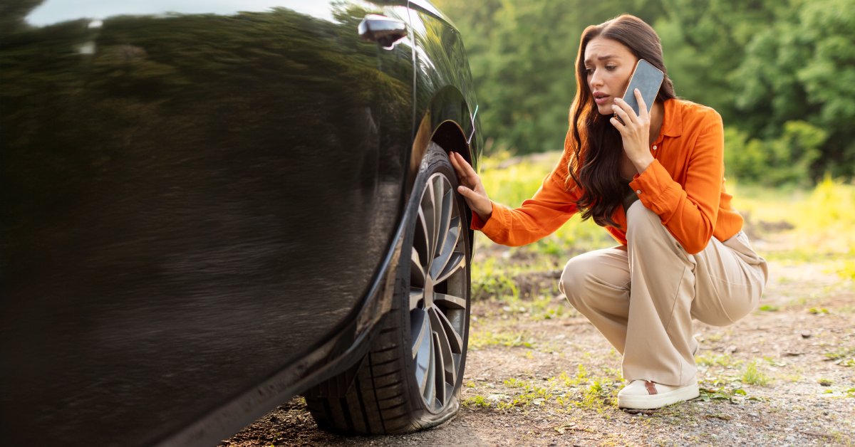 A woman crouched down next to her vehicle while on the phone. She is looking at her flat tire.
