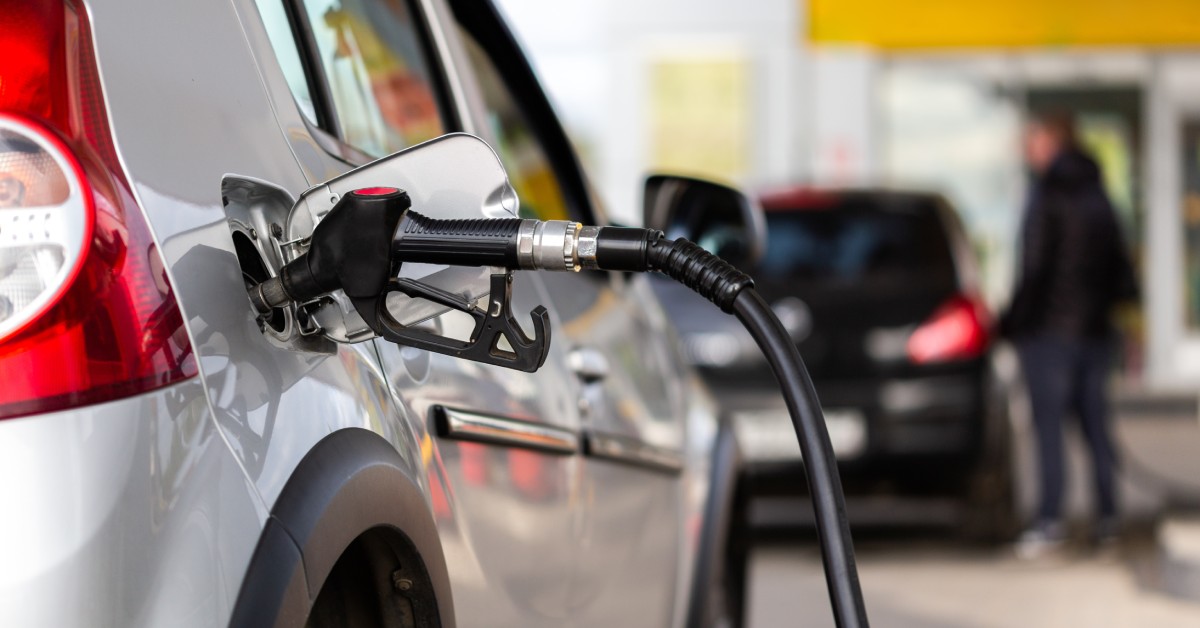 A gray metallic vehicle refueling at a gas station. In the background, there is a blurred car and a person.