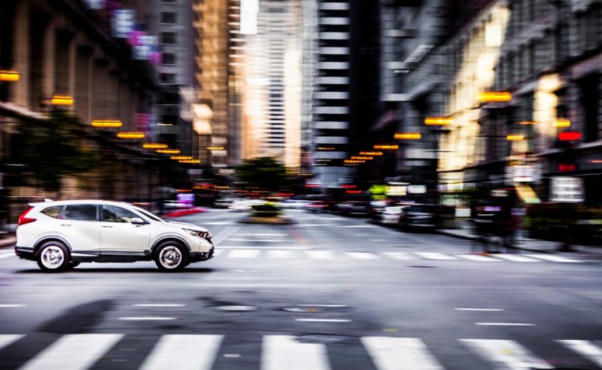 A car driving through an intersection with a crosswalk, pedestrians, streetlights, and tall buildings around it.