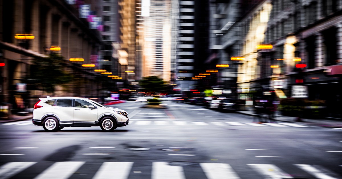 A car driving through an intersection with a crosswalk, pedestrians, streetlights, and tall buildings around it.